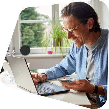 Ein lächelnder Mann, der eine Brille trägt, sitzt an einem Schreibtisch vor einem Laptop und hält ein Smartphone in der Hand