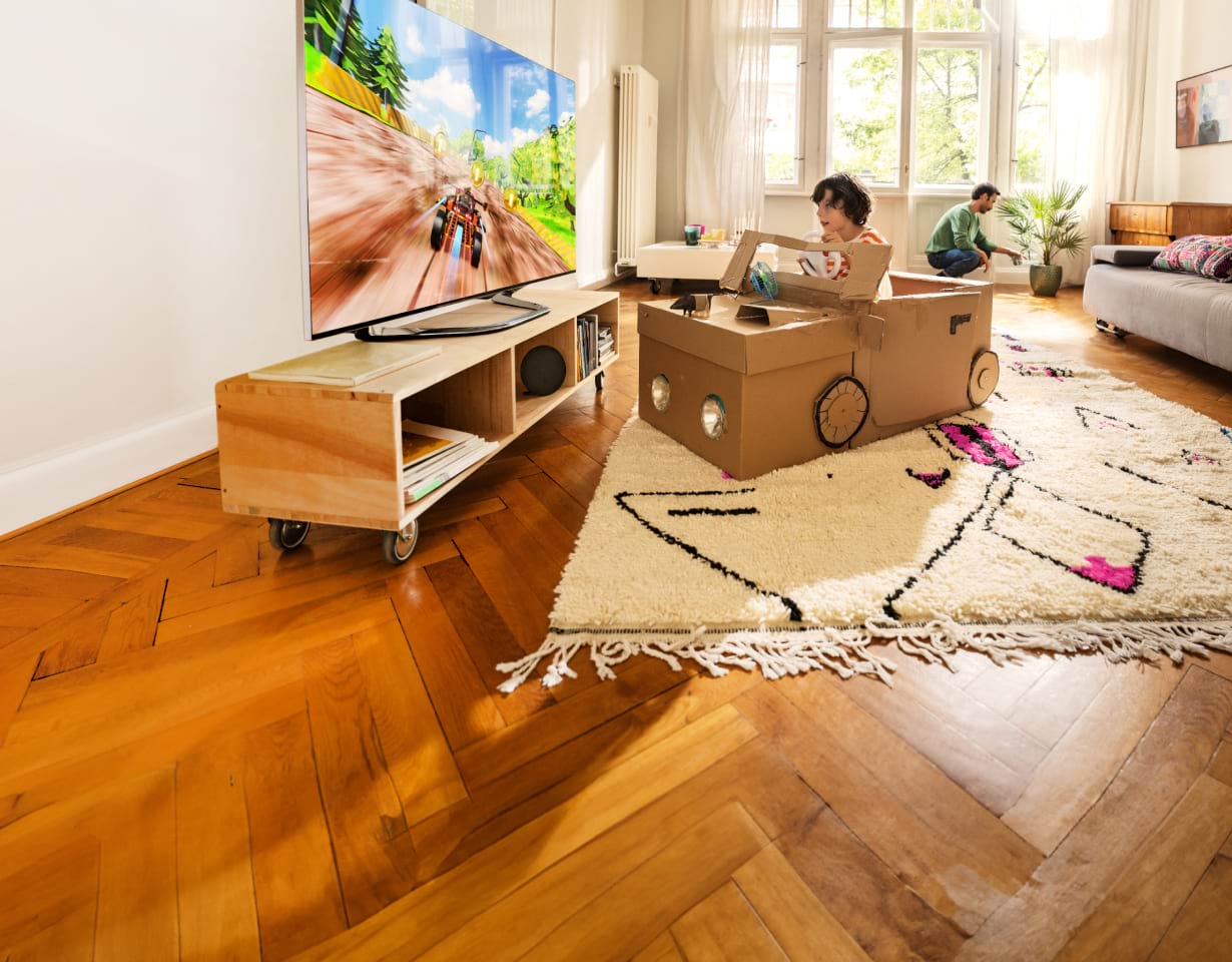 Kleiner Junge der in einem Auto aus Pappkartons sitzt und Autorennen im Fernseher spielt