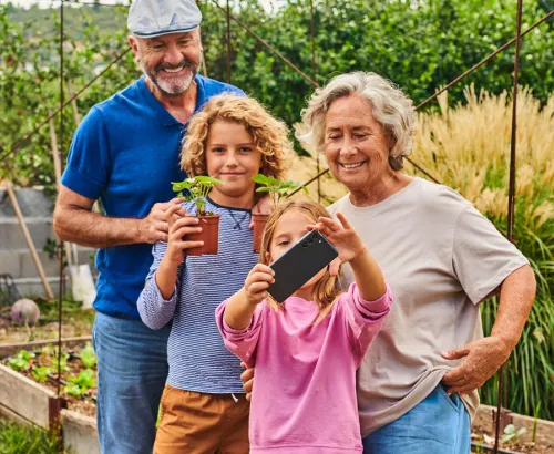 Eine Gruppe von vier Personen in einem Garten. Zwei Personen vorne halten eine kleine Pflanze im Topf hoch. Das Mädchen im Vordergrund hält zudem ein Smartphone in der Hand. Sie sind von Pflanzen und grünem Laub umgeben.