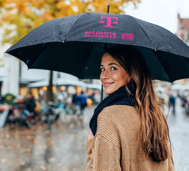 Ein Frau die unter einem Telekom Regenschirm lächelend zurückblickt.