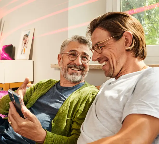 Zwei Männer sitzen lächelnd auf dem Sofa und schauen auf ein Handy