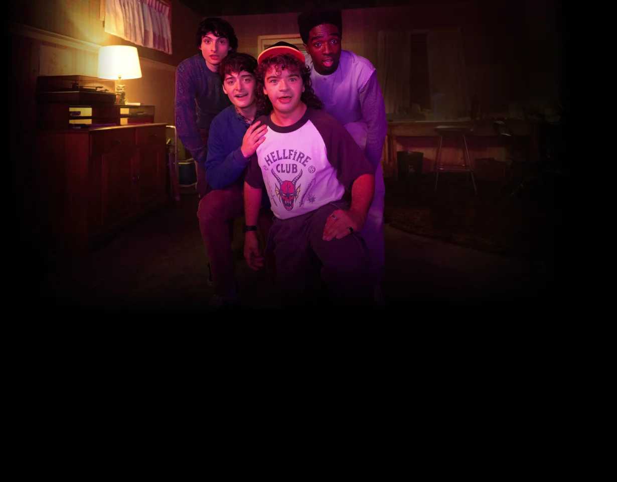 Stranger Things Staffel 5: nur auf Netflix. Jugendliche posieren in einem warm beleuchteten 80er Wohnzimmer mit Lampe und Plattenspieler; vorn ein T Shirt mit der Aufschrift Hellfire Club.