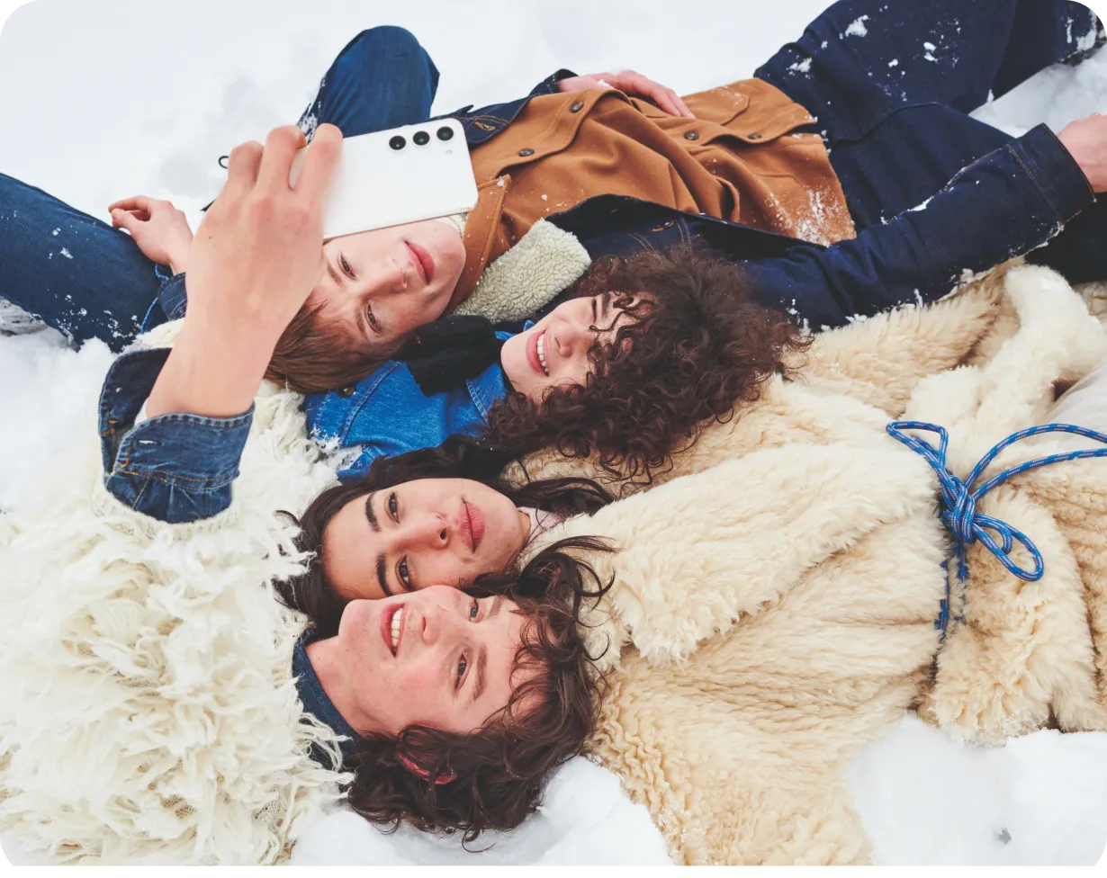 Zwei Päärchen liegen entspannt auf dem Rücken im Schnee und schauen auf ein Smartphones, welches einer von ihnen in hoch hält