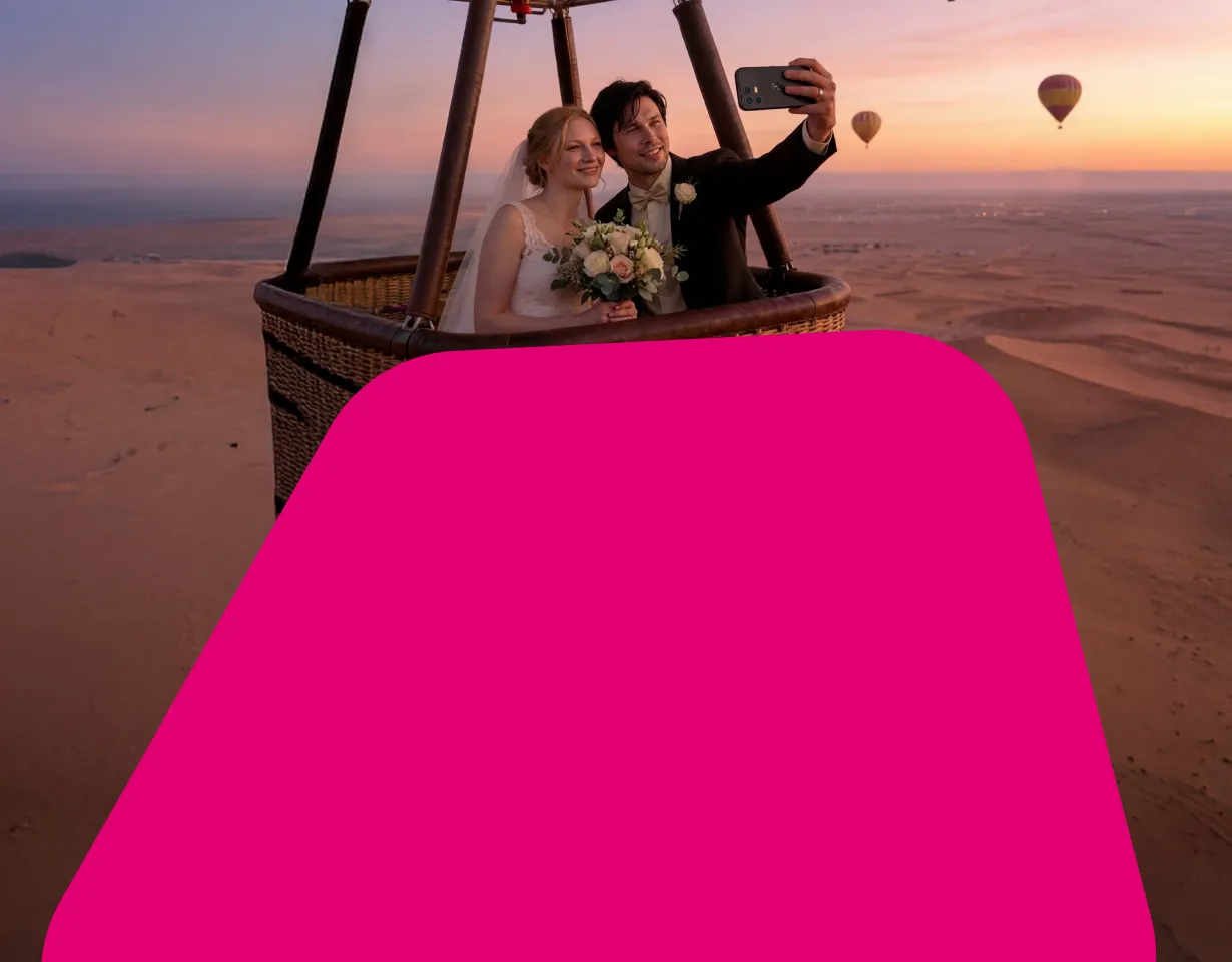 Ein Brautpaar macht ein Selfie in einem Heißluftballon über einer Wüstenlandschaft bei Sonnenuntergang. Die linke Bildhälfte wird von einer großen, abgerundeten magentafarbenen Fläche überdeckt.