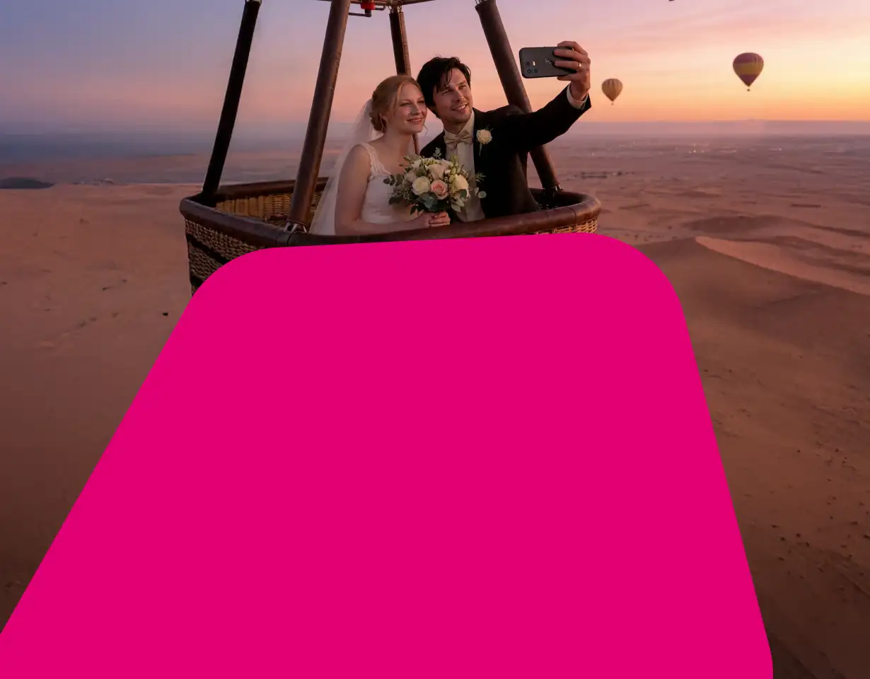 Ein Brautpaar macht ein Selfie in einem Heißluftballon über einer Wüstenlandschaft bei Sonnenuntergang. Die linke Bildhälfte wird von einer großen, abgerundeten magentafarbenen Fläche überdeckt.