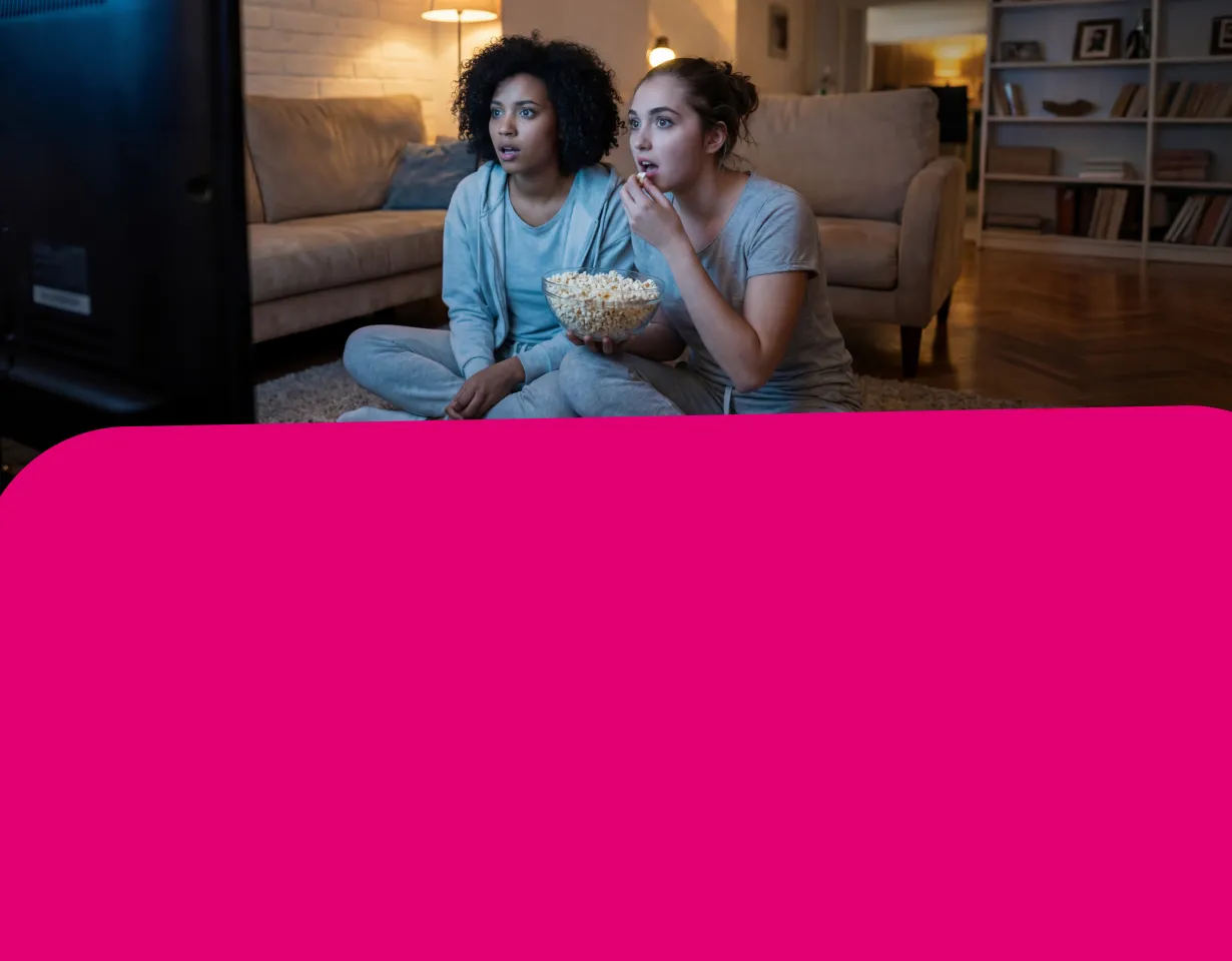 Zwei junge Frauen sitzen abends in einem gemütlichen Wohnzimmer auf dem Boden, essen Popcorn und schauen gespannt in Richtung eines Fernsehers.
