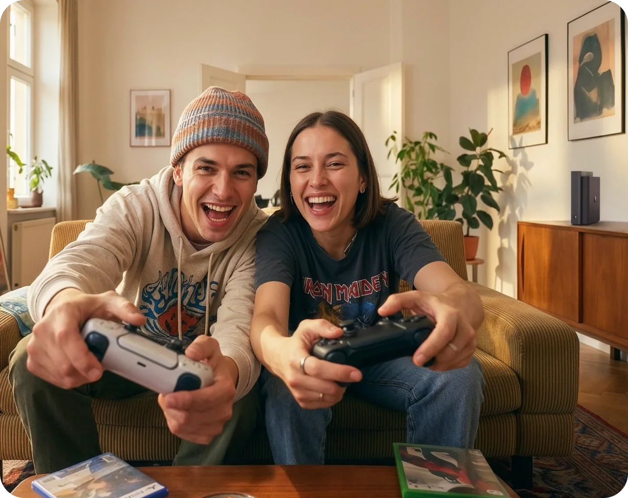 Zwei junge Menschen sitzen lachend auf einem Sofa und spielen gemeinsam Videospiele mit Controllern.