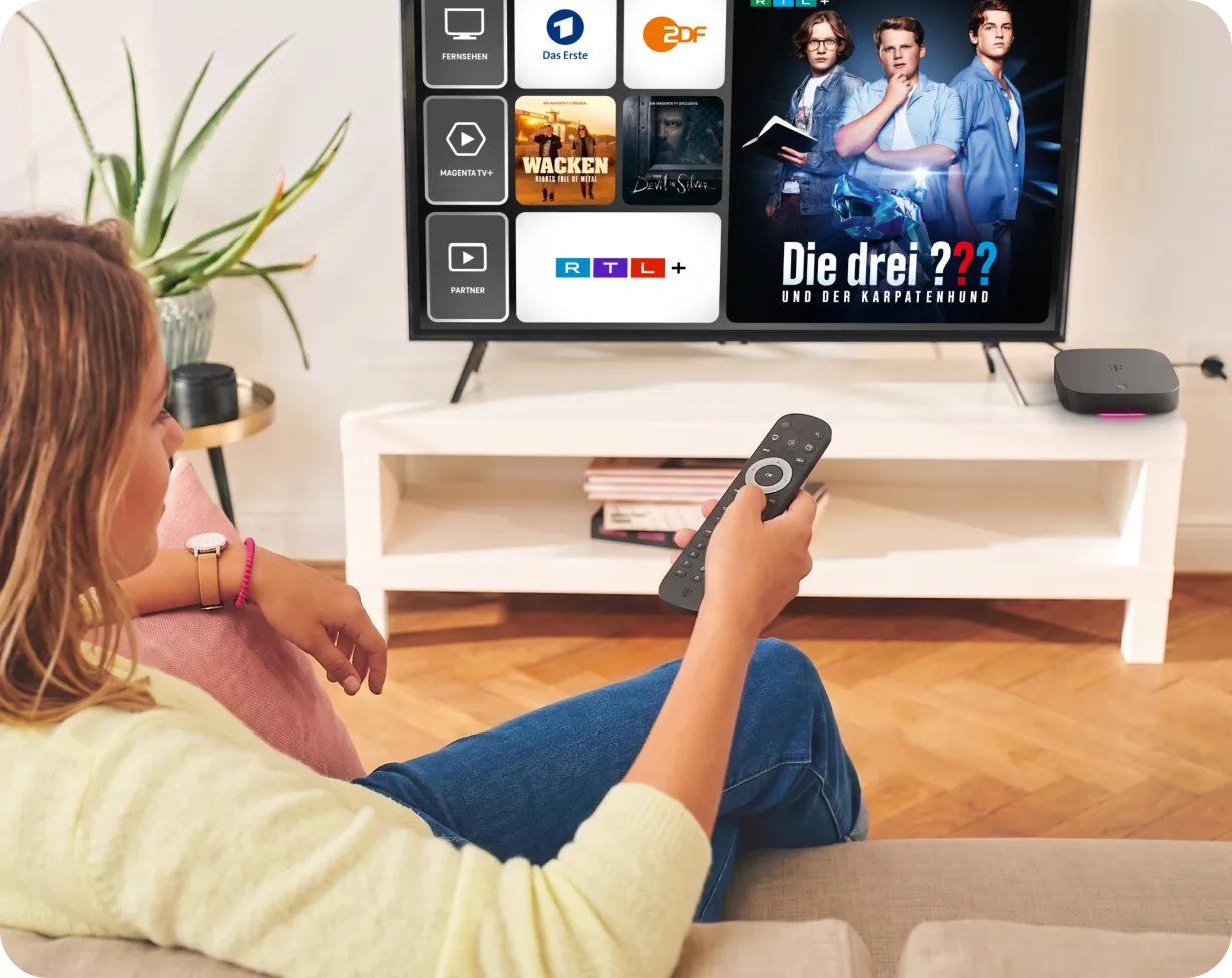 Frau mit Fernbedienung auf dem Sofa vor einem Fernseher mit der MagentaTV Oberfläche: Das Erste, ZDF und RTL+ sowie die Serie Die drei ??? und der Karpatenhund